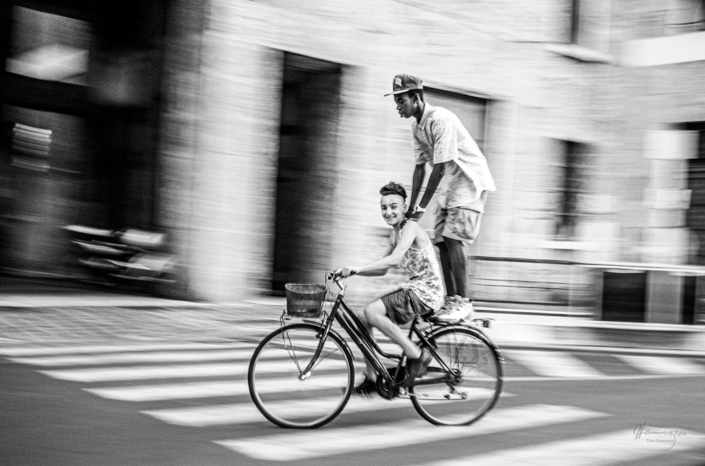 Panning photograph of two boys riding a bicycle through the streets of Brescia, Italy at evening, the background blurred by camera movement into streaks of warm light.