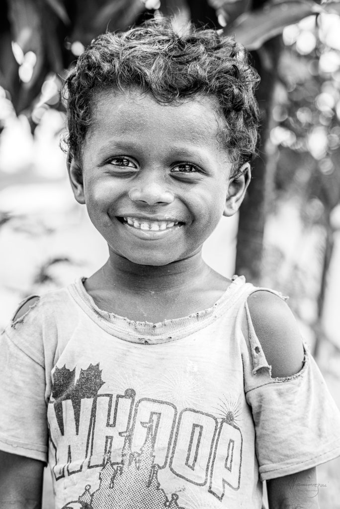 Portrait of a young boy in a torn t-shirt, laughing directly at the camera with complete, unguarded joy, photographed in Nepal.