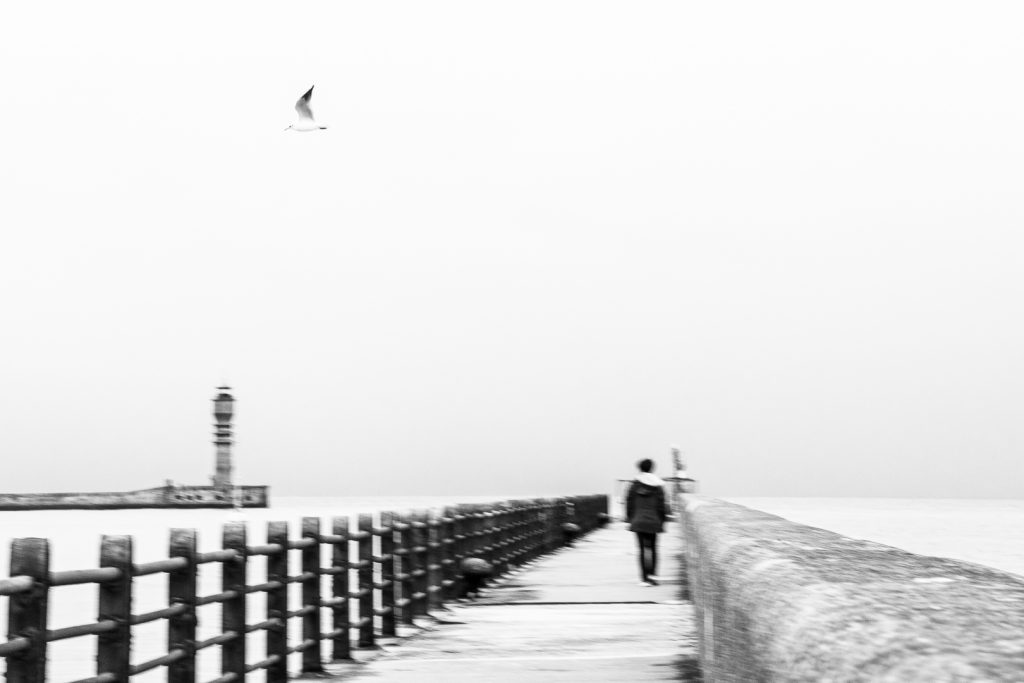 Black and white photograph of a solitary figure walking away along a misty pier in Dunkirk, France, a seagull suspended in the pale sky above.