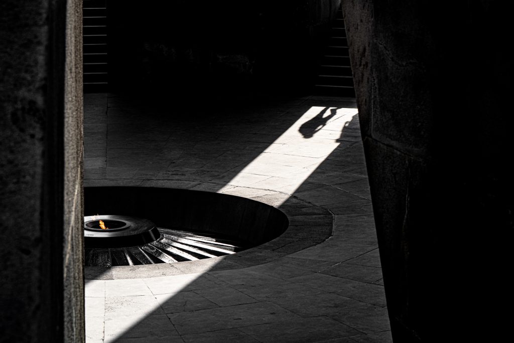 Black and white photograph of the eternal flame at the Armenian Genocide Memorial in Yerevan, a small orange flame burning in a circular stone basin, framed by a dramatic shaft of light cutting across the dark stone floor, with the silhouetted shadows of two departing visitors cast across the light.