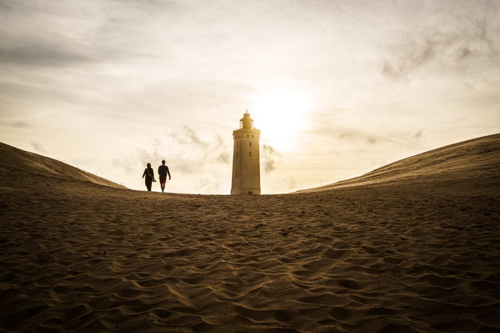 Two silhouetted figures walking across sand dunes toward the Rubjerg Knude lighthouse at sunset, the lighthouse backlit by a warm glowing sky, the sweeping dunes curving on either side.