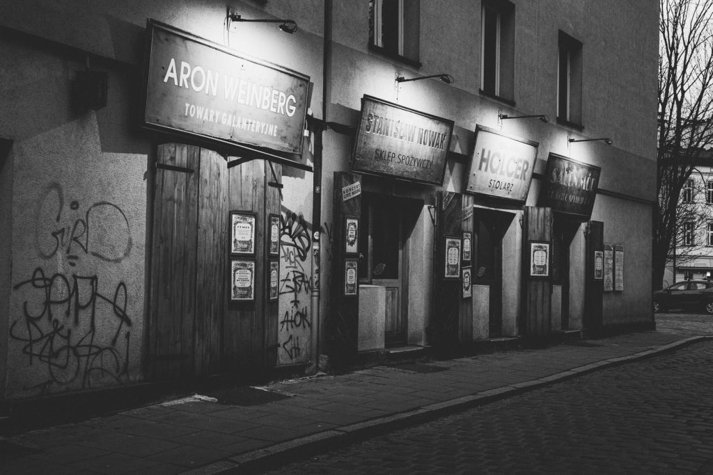 Black and white night photograph of a row of reconstructed pre-war Jewish shopfronts in the Kazimierz district of Kraków, Poland, their illuminated signs bearing names including Aron Weinberg and Stanisław Nowak, the wooden doors covered in graffiti, the street empty and quiet.