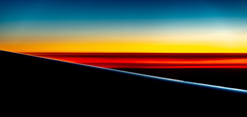 Aerial view from airplane window showing wing silhouette against layered sunset sky in shades of orange, gold and deep blue.