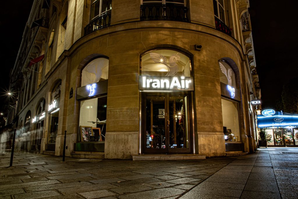 Night photograph of the IranAir office on the Champs-Élysées in Paris, its illuminated sign and blue neon Huma bird logos glowing against the warm stone of a curved Haussmann building on an empty pavement, the street quiet and dark around it.